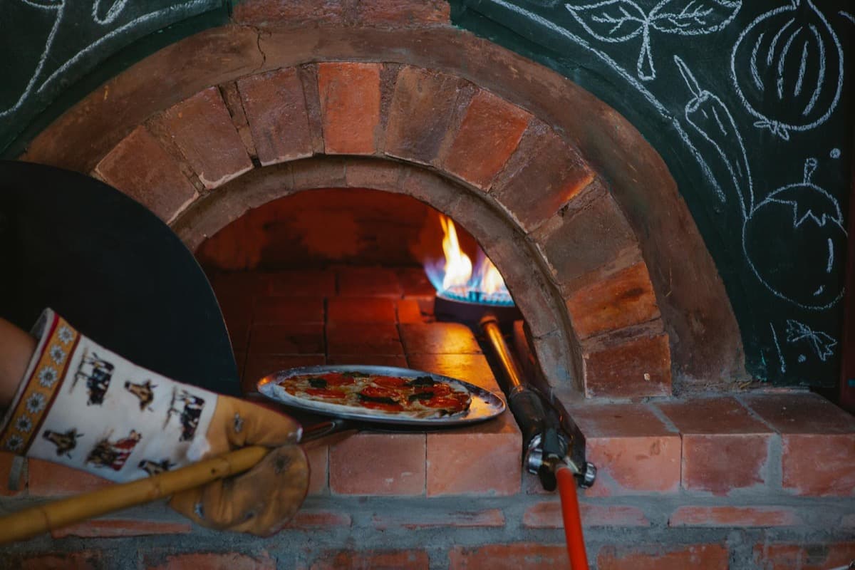 A pizza being pulled from a flaming wood-fired brick oven (16:9)