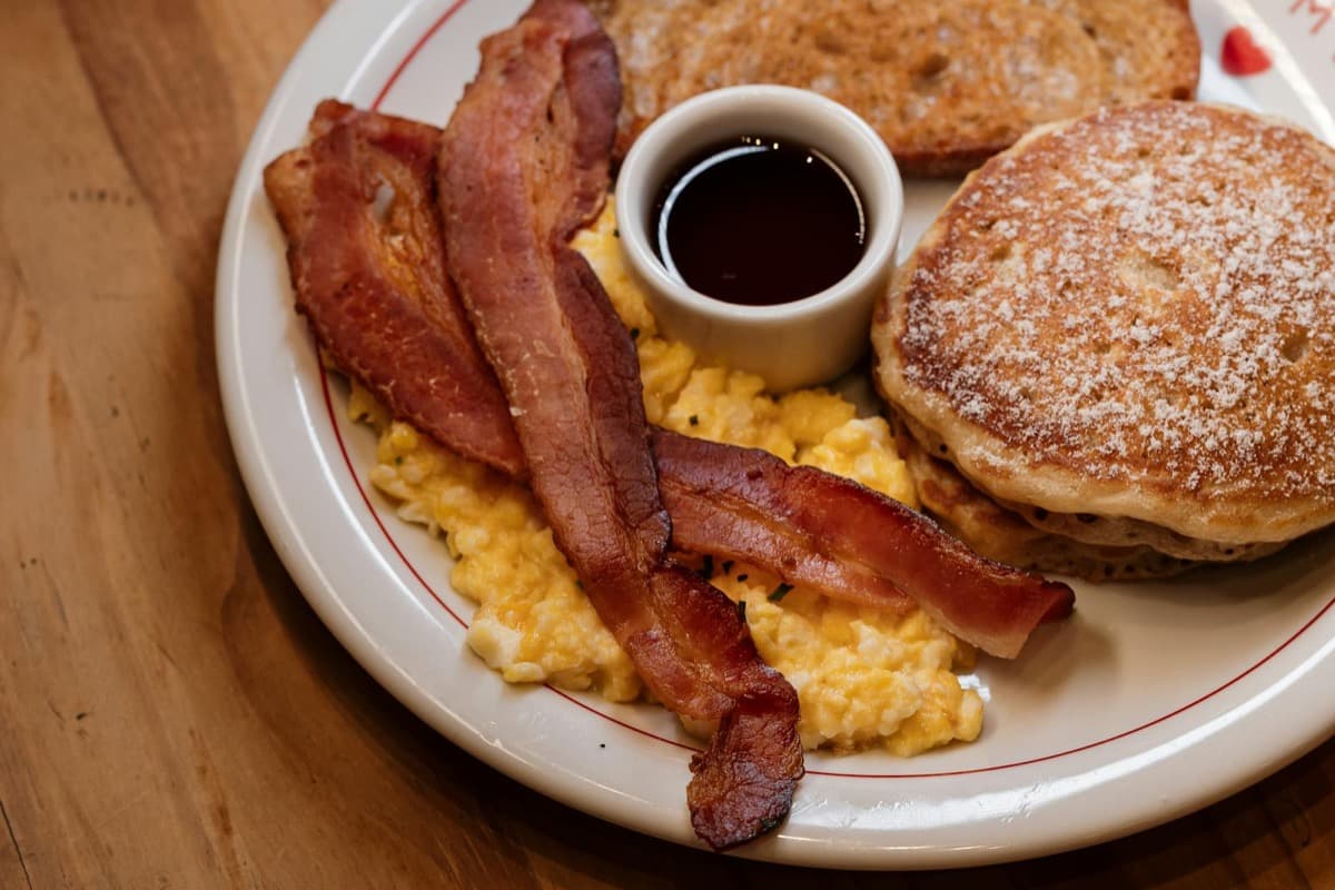 A plate of pancakes, crispy bacon, and scrambled eggs with a cup of syrup (16:9)