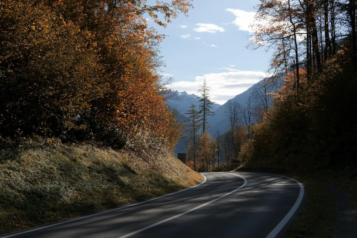 A two-lane road curving through autumn forest toward distant mountains (16:9)