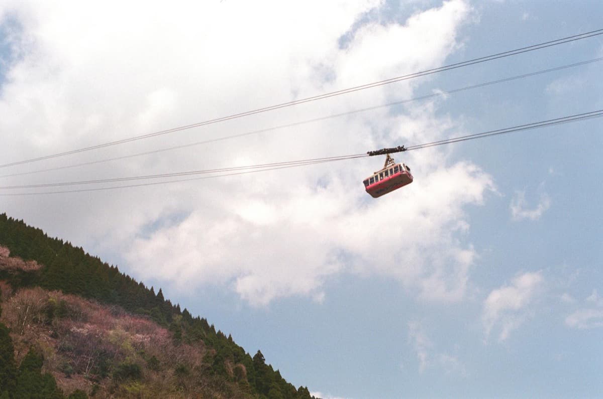 A red aerial tramway car gliding on cables above a forested mountain slope (16:9)