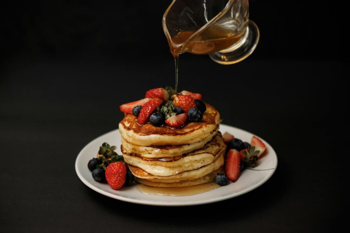 Syrup being poured over a tall stack of pancakes topped with strawberries and blueberries (16:9)