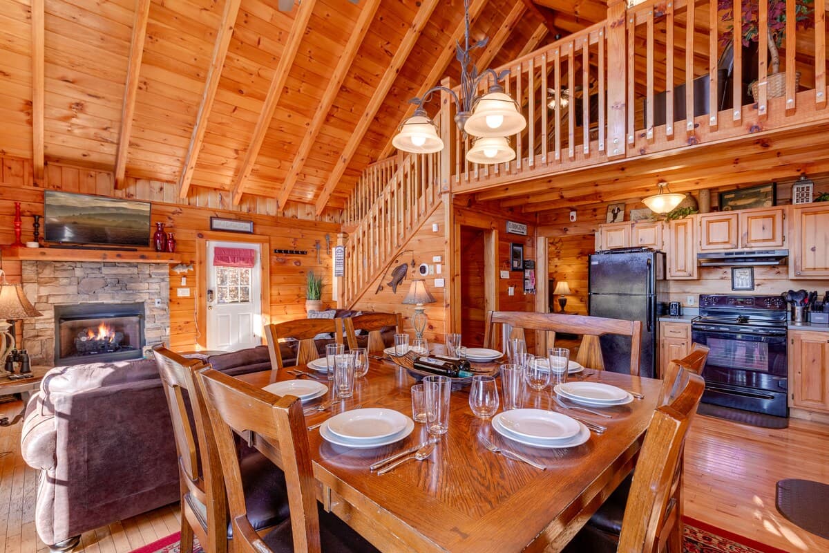 Dining room table set for six with stone fireplace, kitchen, and stairs to loft visible
