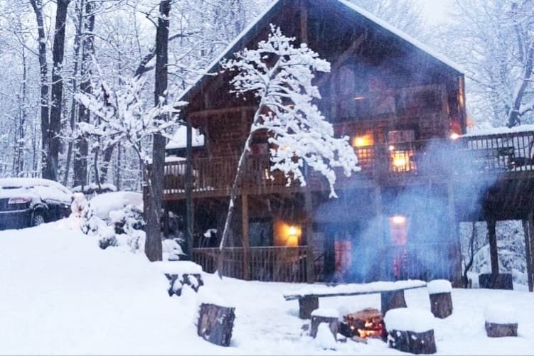 Cabin glowing in a winter wonderland with heavy snow covering the fire pit, log benches, and trees