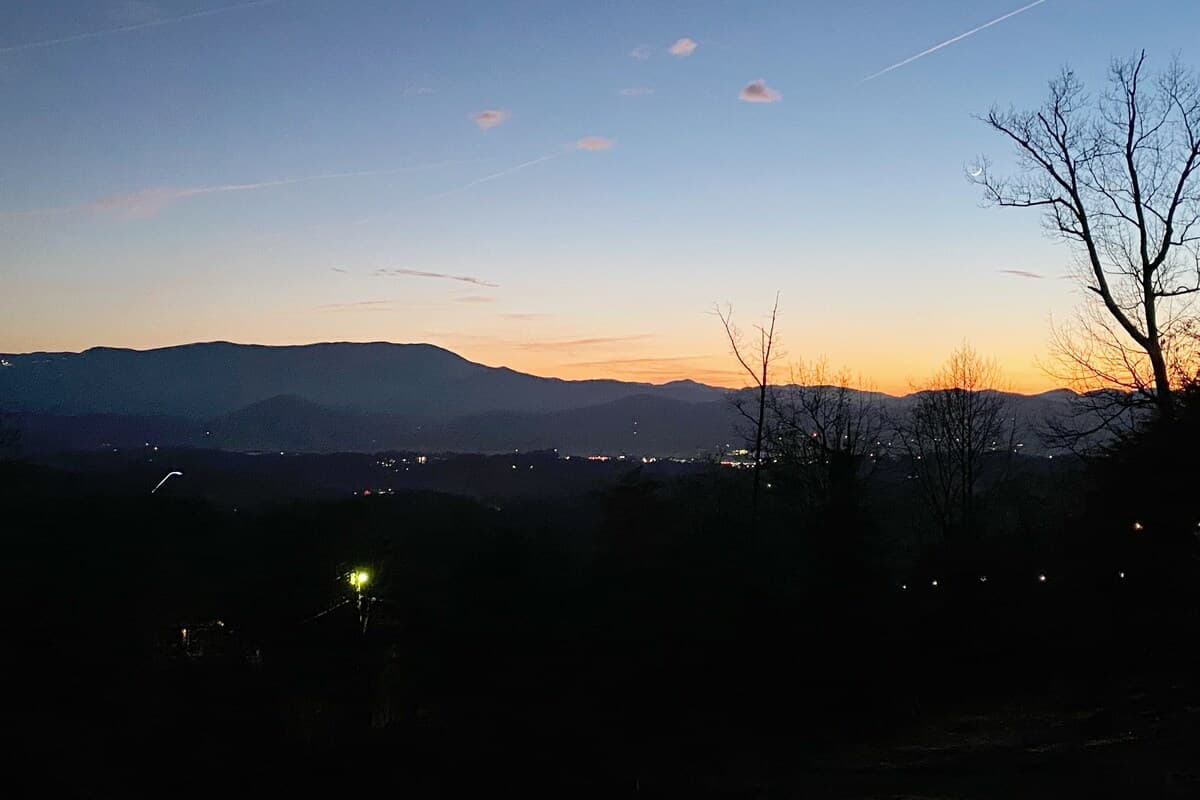 Sunset silhouetting Smoky Mountain ridges with crescent moon and city lights below
